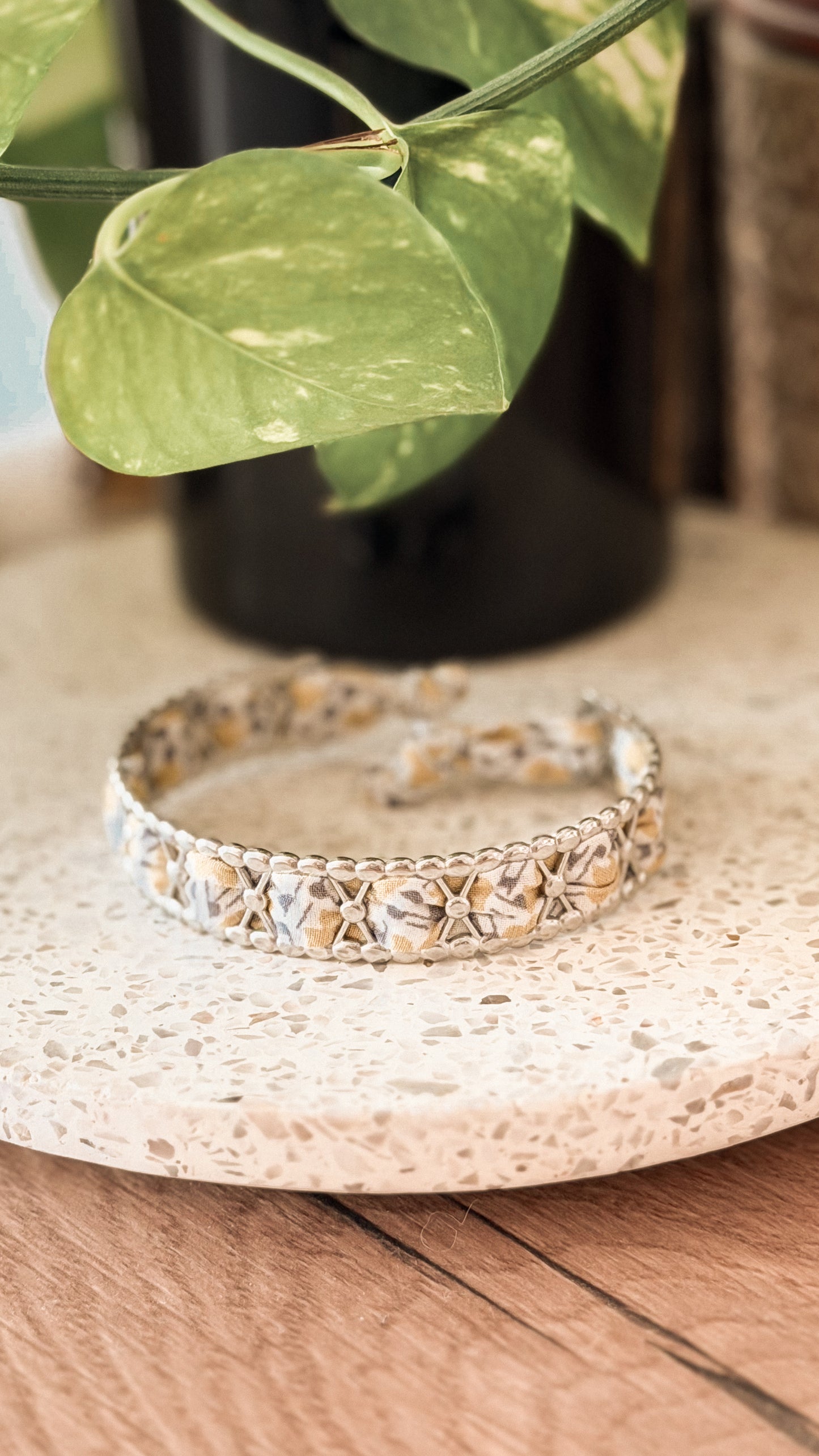 Bracelet jonc en acier inoxydable et tissu upcyclé argenté motif feuilles beige, posé sur un plateau en terrazzo